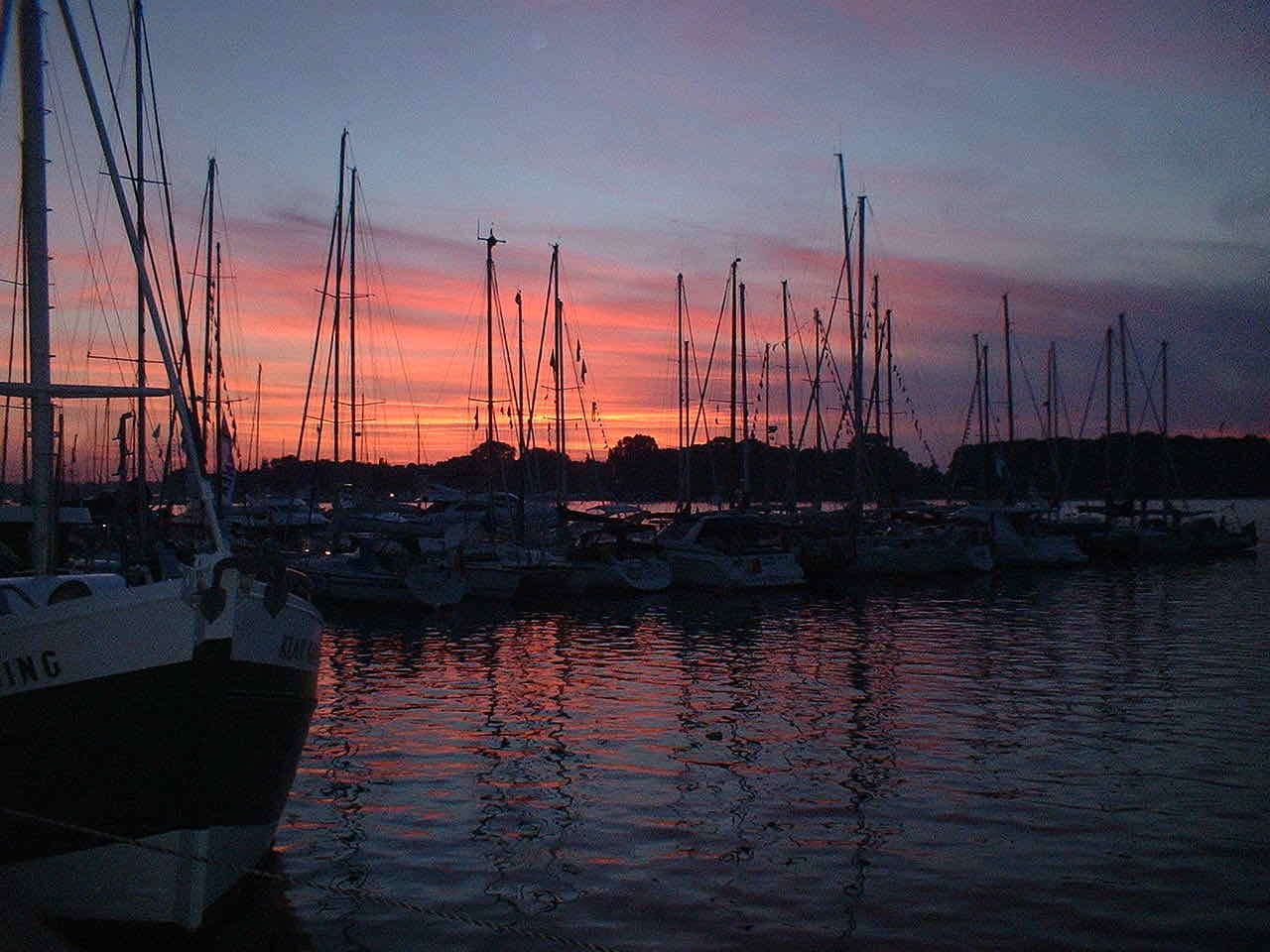 Masts of moored sailing boats in Rostock harbour against a red and purple evening sky, with the bow of a dark ship at the left
