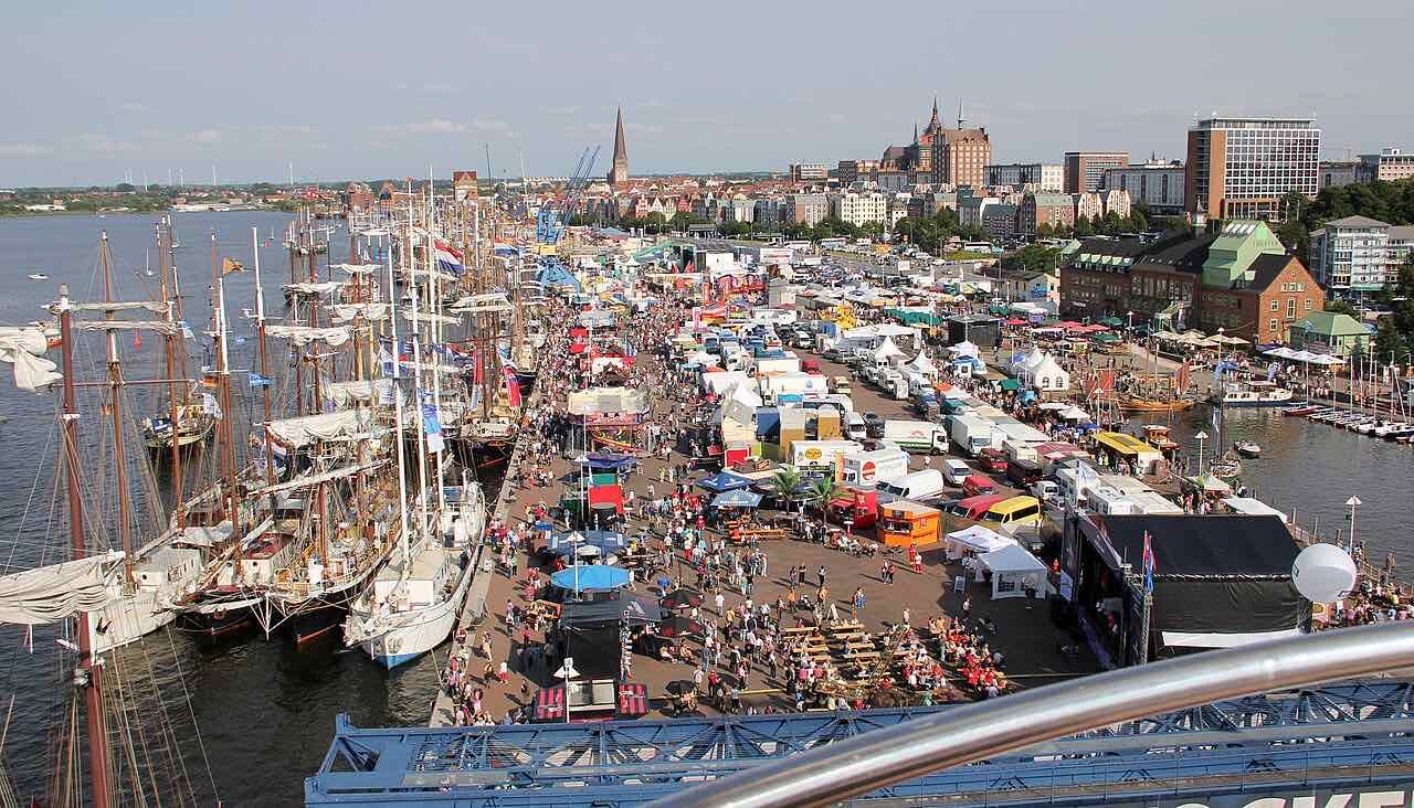 Looking down on the Rostock harbour during Hanse Sail, with tall ships along the quay, market tents and crowds, and church spires in the distance