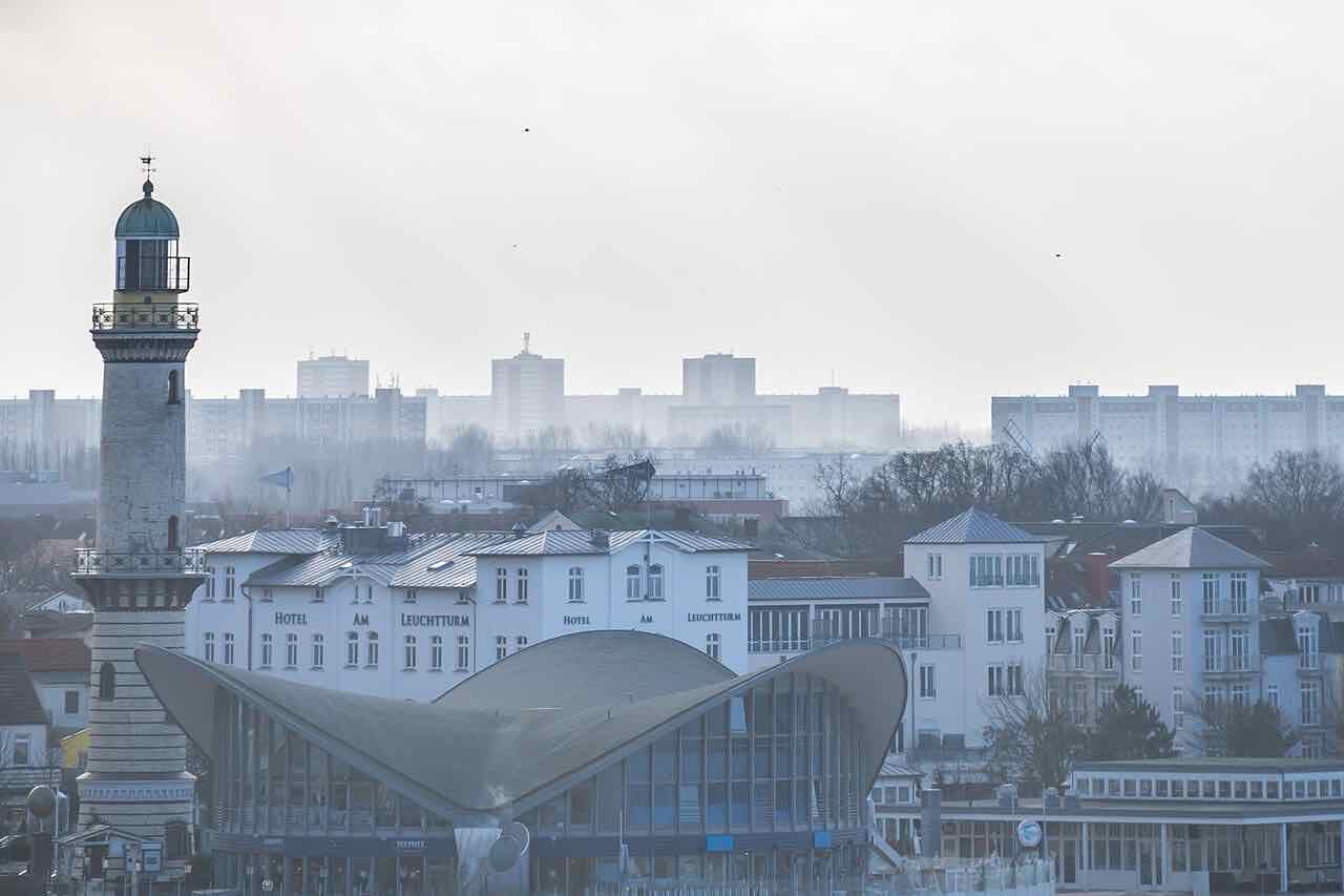 The Warnemünde lighthouse tower beside the curved-roof Teepott building, with white hotels and grey housing blocks of Rostock behind in haze