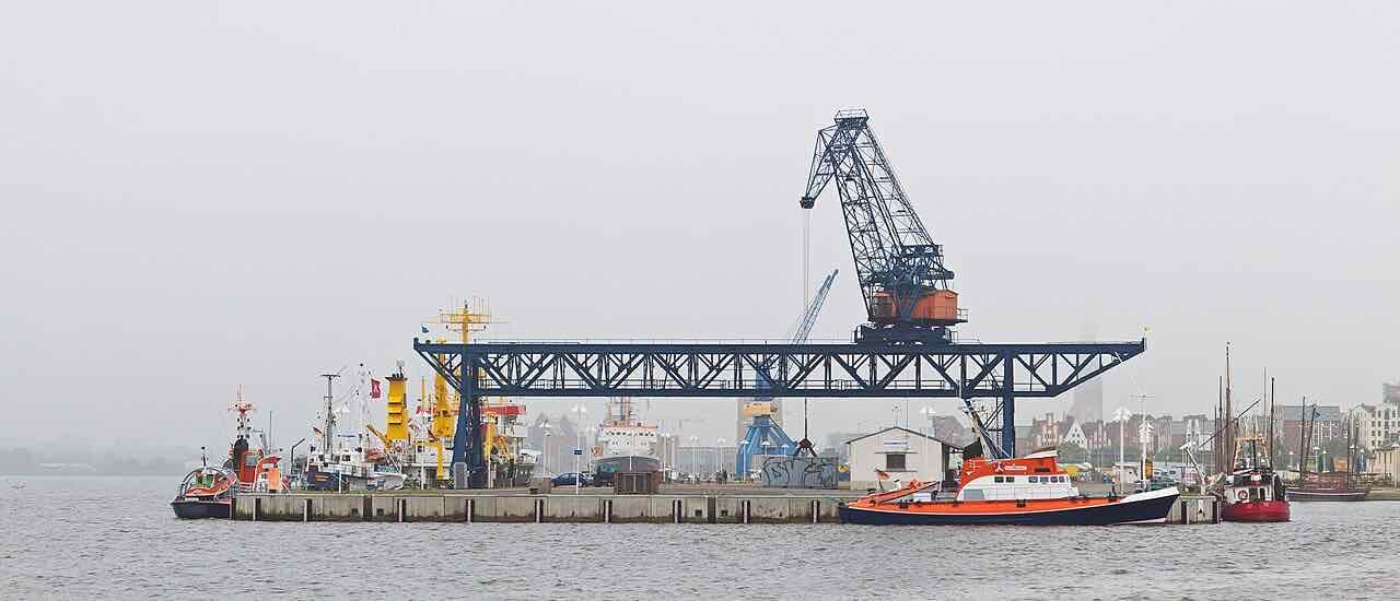 A pier in the Rostock city harbour with a blue gantry crane, lifeboats and survey ships, and old sailing boats and houses on the far side under a grey sky