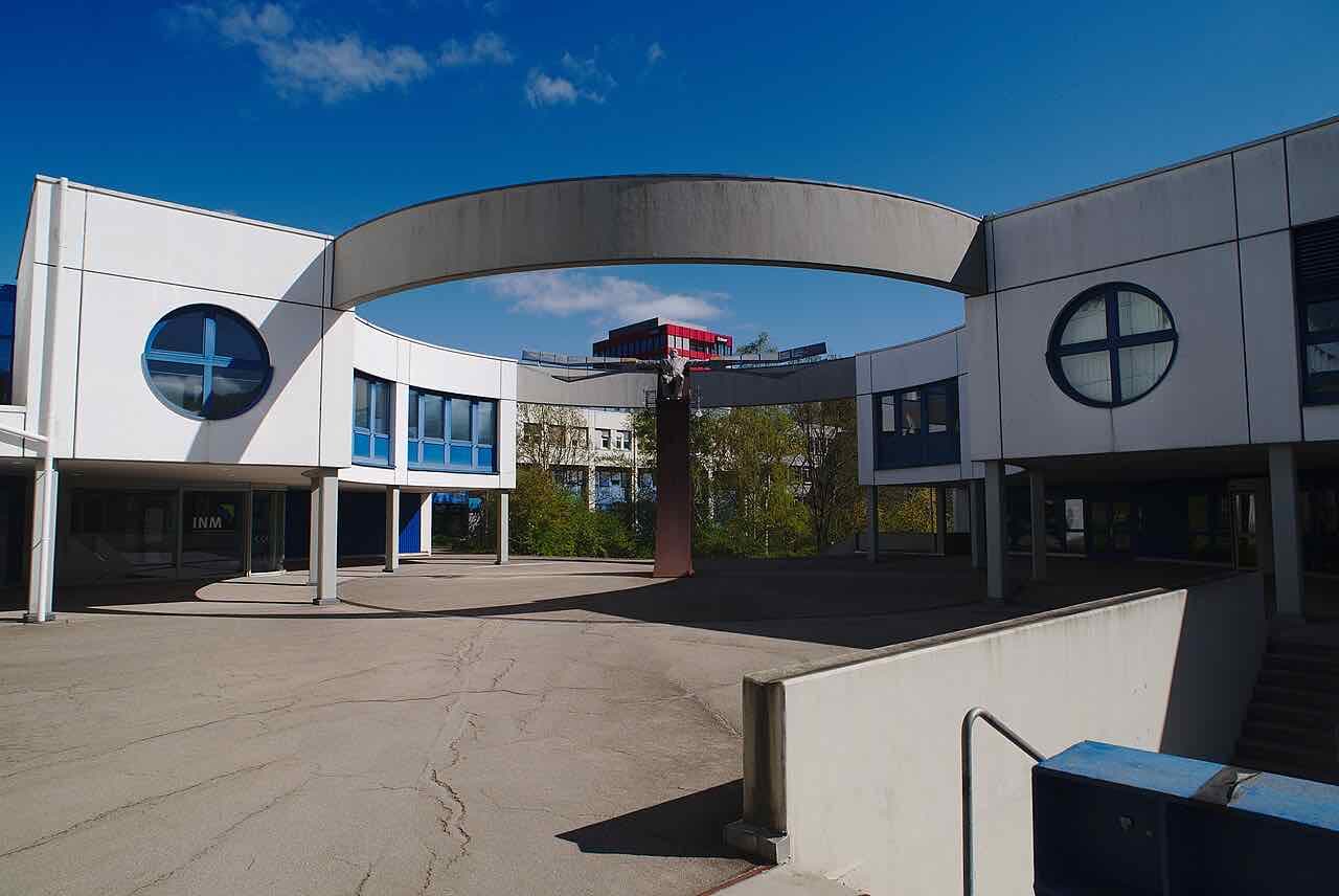 White campus buildings of Saarland University joined by a curved walkway, with round blue-framed windows and a blue sky