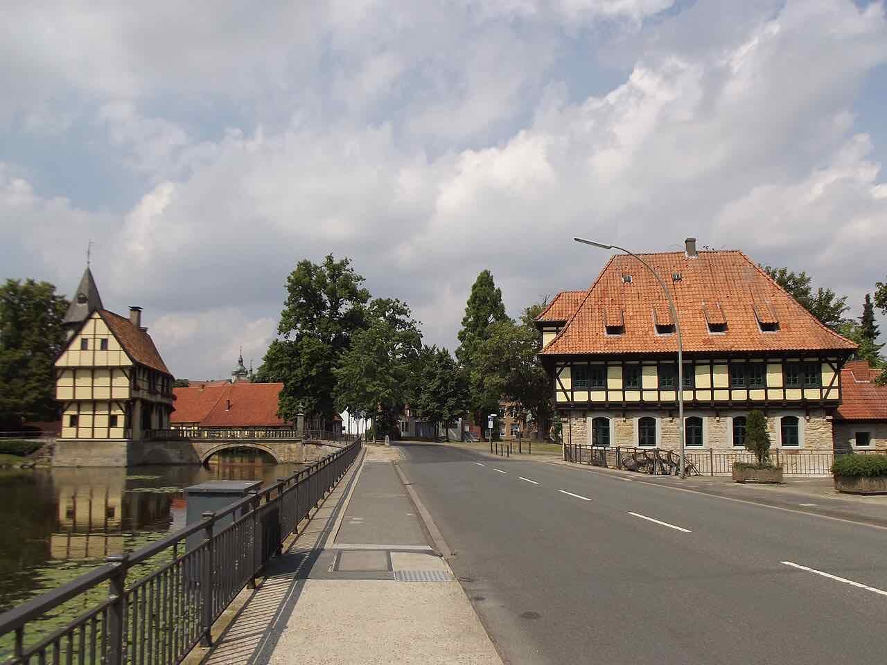 A half-timbered gatehouse with a small spire on the left beside a stone bridge over still water, and on the right a large half-timbered mill with a steep red tiled roof beside an empty road under a cloudy sky