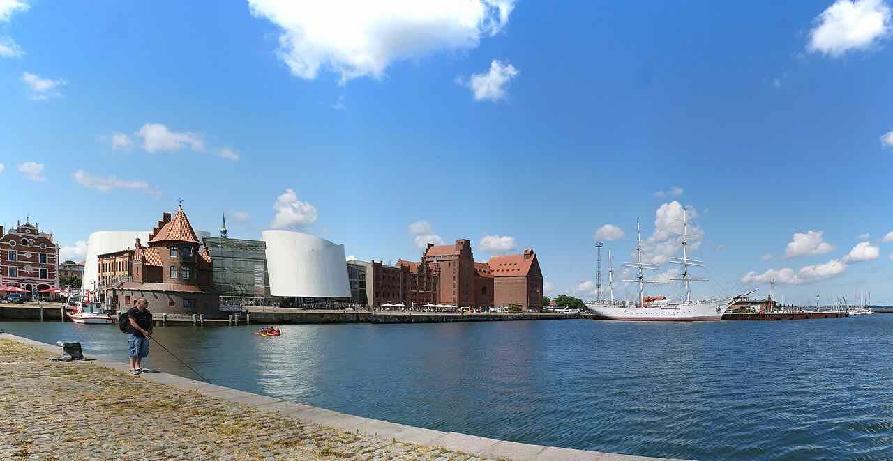The Stralsund harbour on a sunny day, with the white curved Ozeaneum, red-brick warehouses and the three-masted sailing ship Gorch Fock I moored on the right