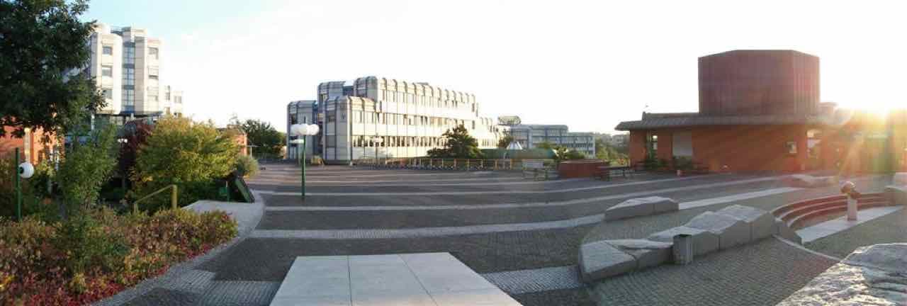 A wide stone-paved square on the Trier University campus with concrete faculty buildings and a low sun