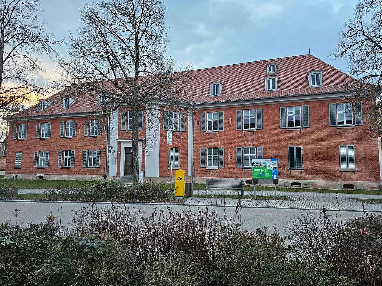 The Rotes Schloss in Triesdorf, a long two-storey red-brick building with grey shutters, a steep tiled roof with dormers and a pale central entrance, seen across a road at dusk