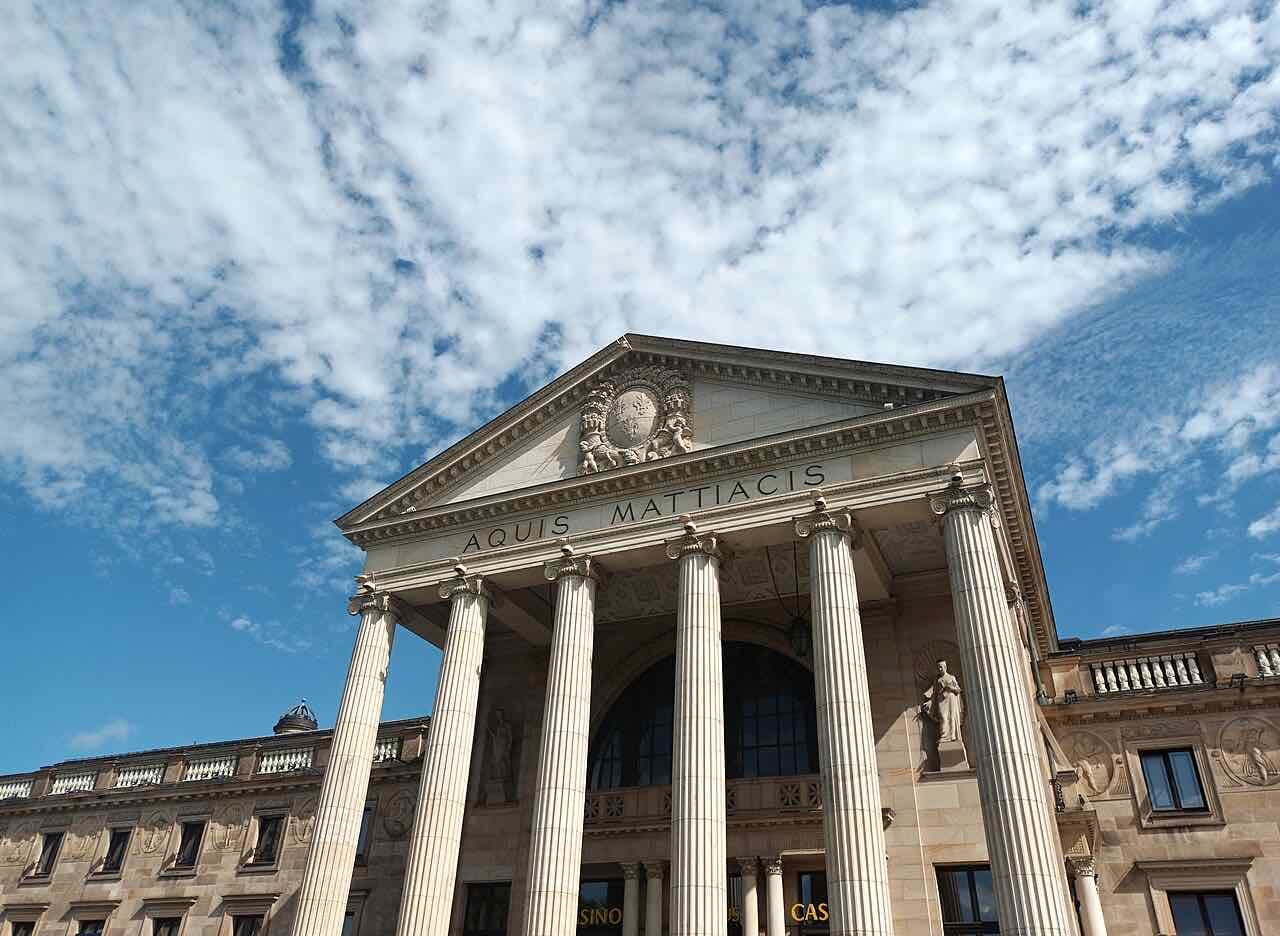 The columned entrance of the Wiesbaden Kurhaus under a blue sky with scattered clouds, with the Latin words AQUIS MATTIACIS carved over the columns