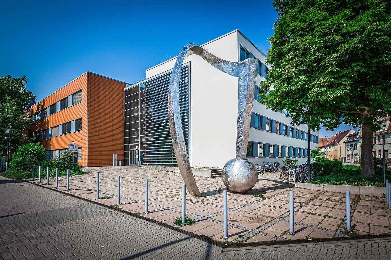 Building N of Hochschule Worms, with an orange and a white facade, a large silver sculpture of a curved arch on a ball and bicycles parked beside it