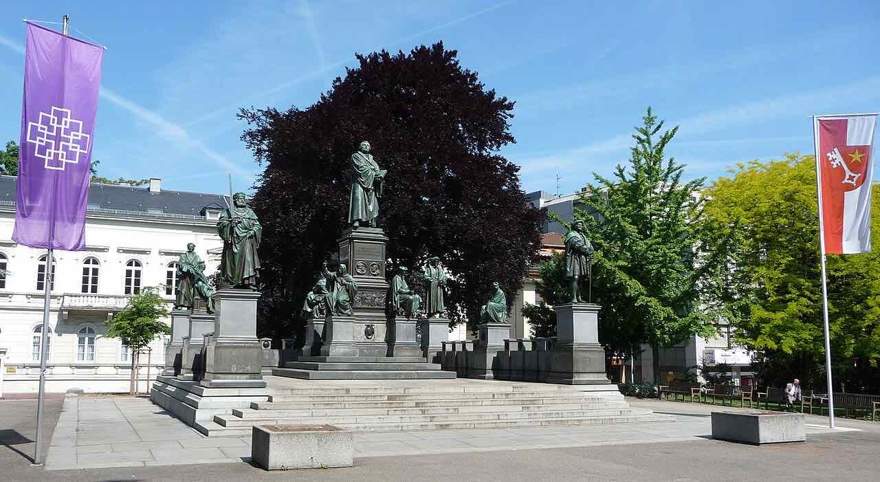 The Luther Monument in Worms: a tall bronze Luther on a stepped stone base, surrounded by smaller bronze figures, with a dark copper beech and a Worms city flag beside it