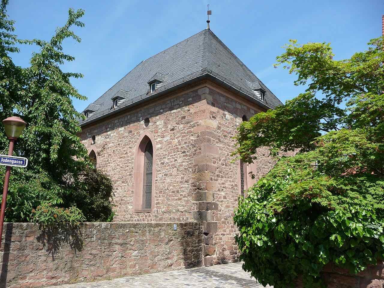 A red sandstone building with a steep slate roof and tall pointed windows beside a street sign reading Judengasse, framed by green trees