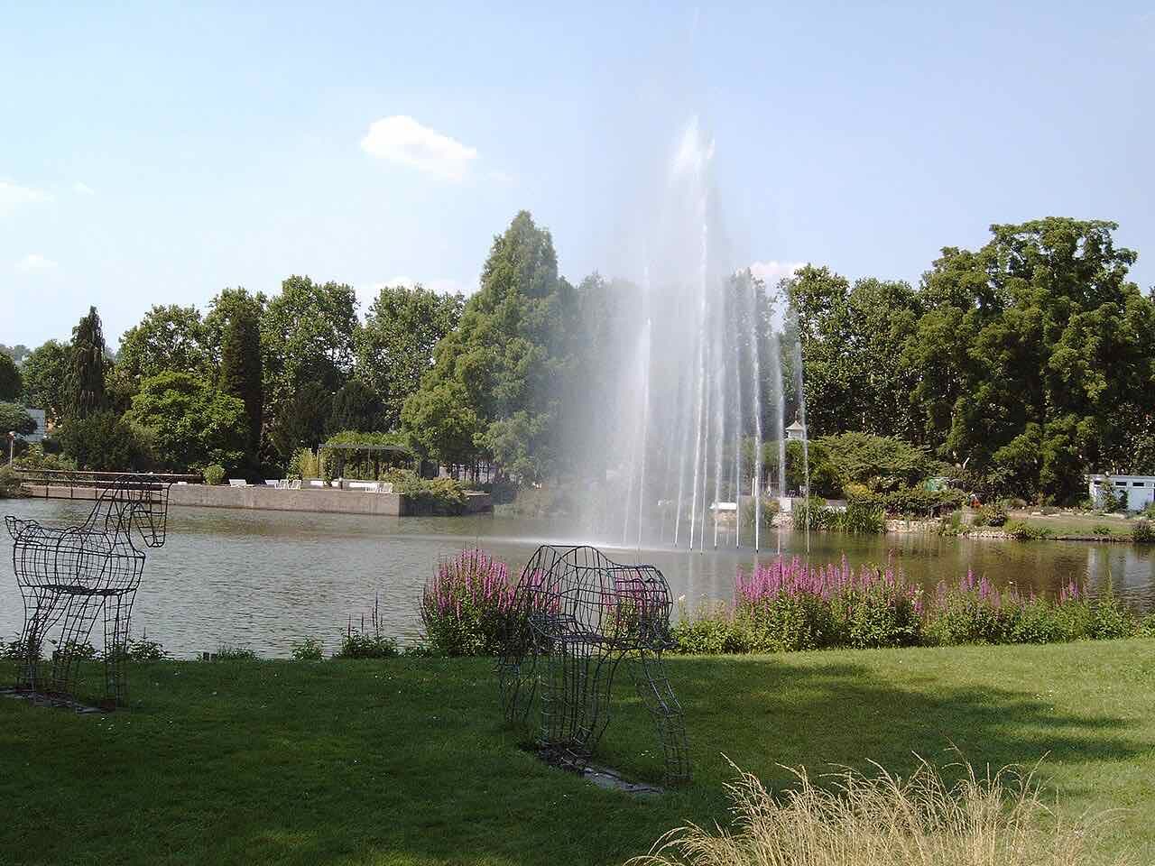 A pond with a tall fountain in the middle of the Zweibrücken rose garden, with purple flowers on the bank, wire sculptures of animals on the lawn and tall trees behind