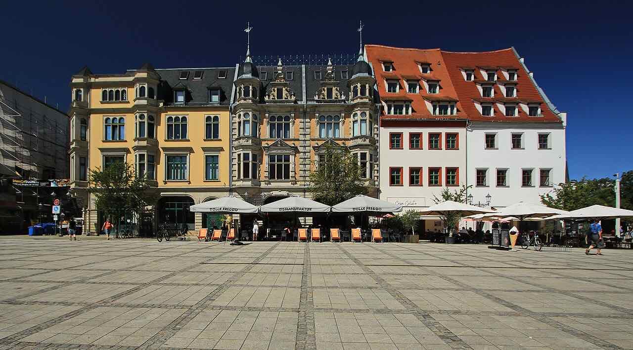 A wide cobbled market square under a deep blue sky with café umbrellas and orange deck chairs along its edge, and behind them a row of tall houses: a yellow one, an ornate pale one with small corner turrets and spires, and a white one with a steep red tiled roof